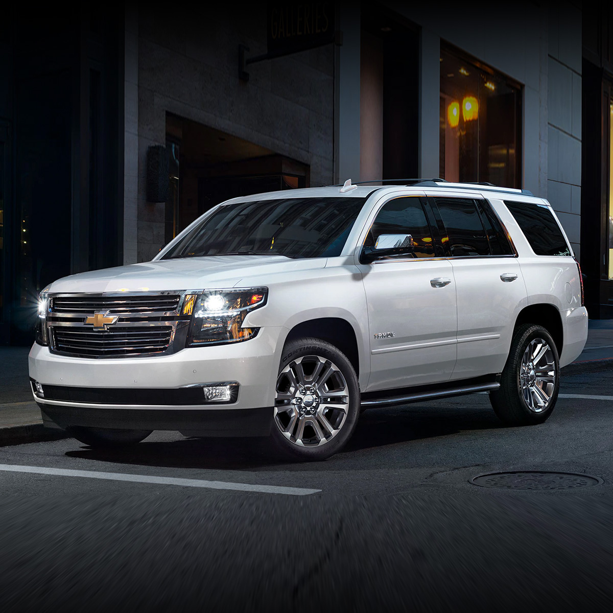 side view of white chevrolet tahoe suv parked in front of a building with headlights turned on