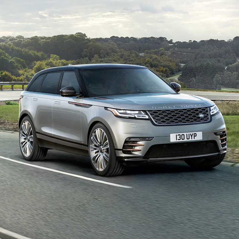 frontal profile of blue land rover range rover velar suv accelerating on an asphalt road with green trees around it