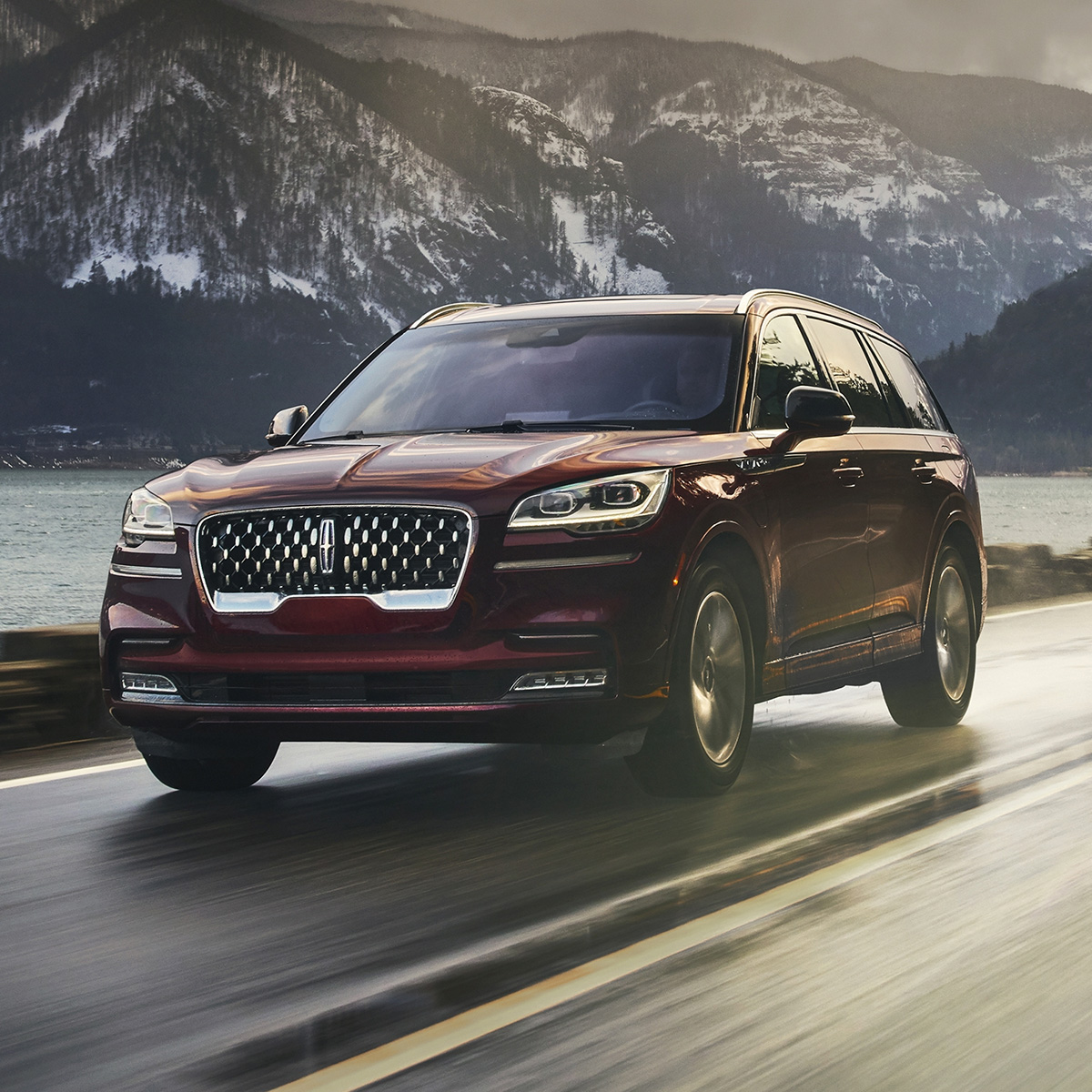 side profile of dark red lincoln aviator suv accelerating on a road that is next to a lake sorrounded by mountains