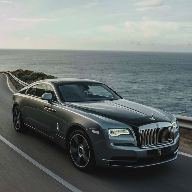 frontal profile of black Rolls Royce wraith accelerating on a road next to open water