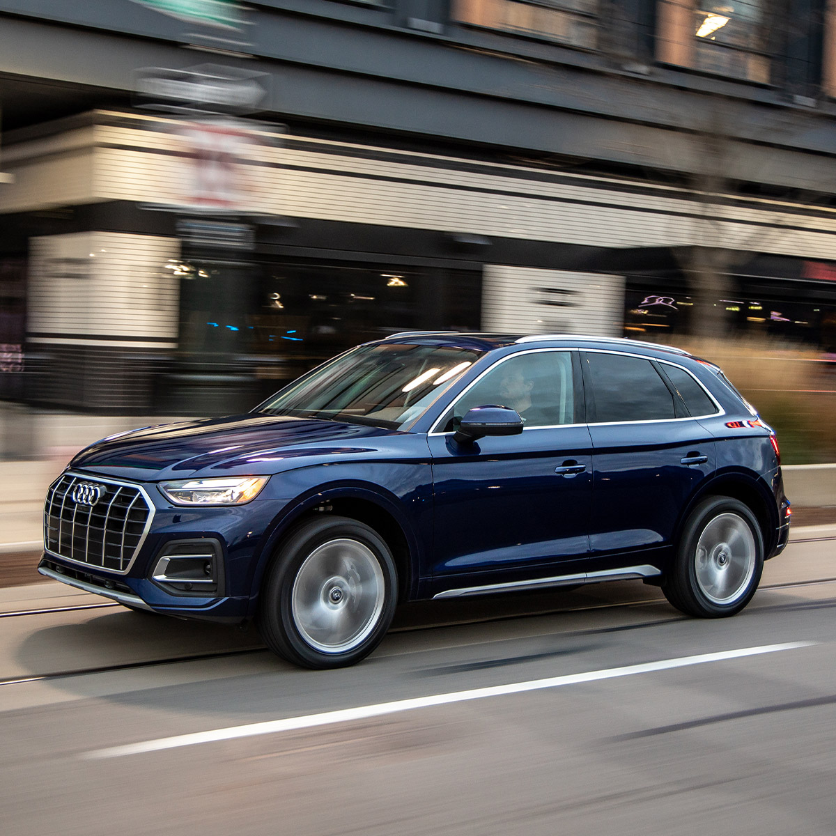 side profile of audi Q5 suv in dark blue color driving down a city street in front of a commercial building
